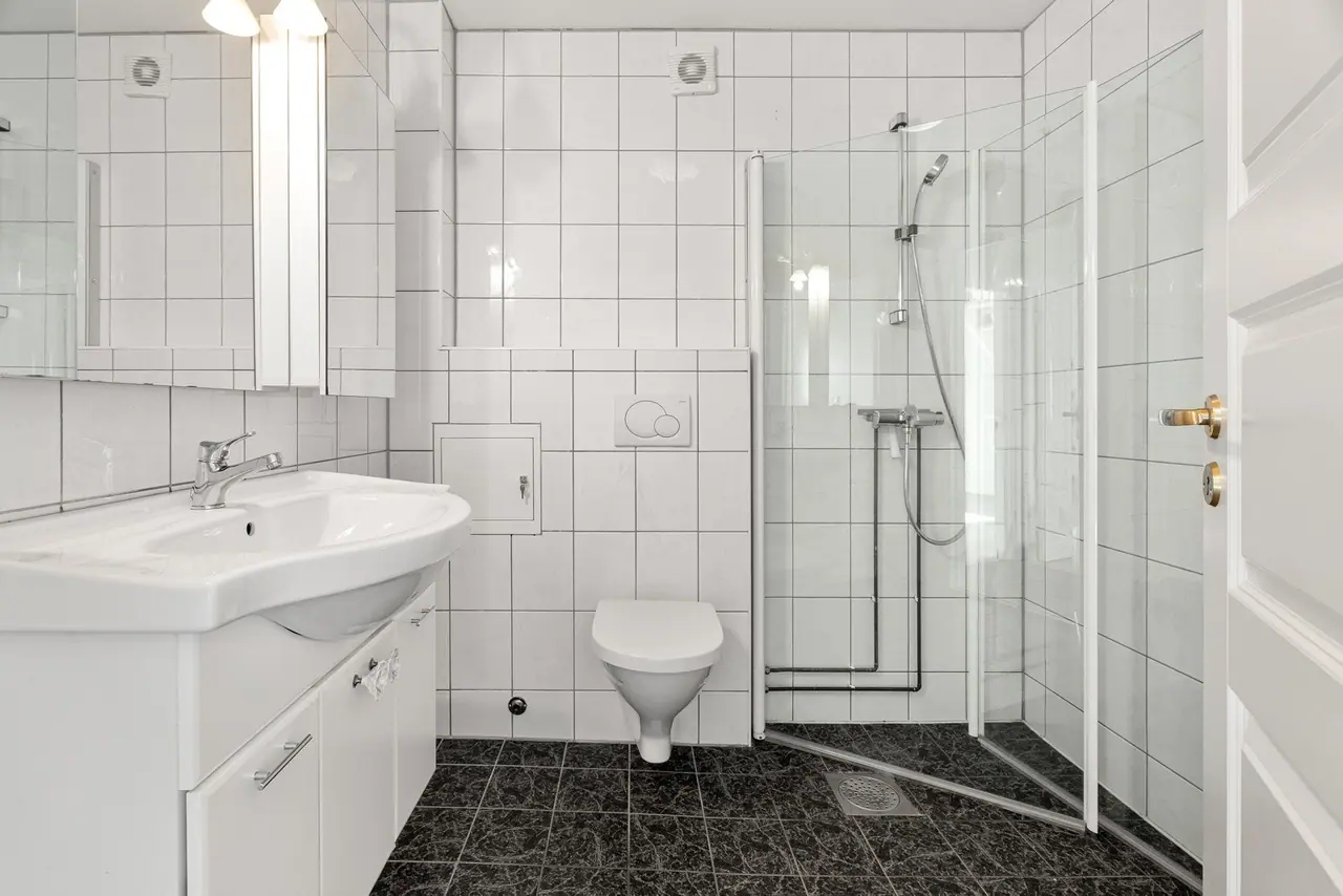 Lyst flisbelagt bad med hjørnedusj med glassdører, toalett og servant.